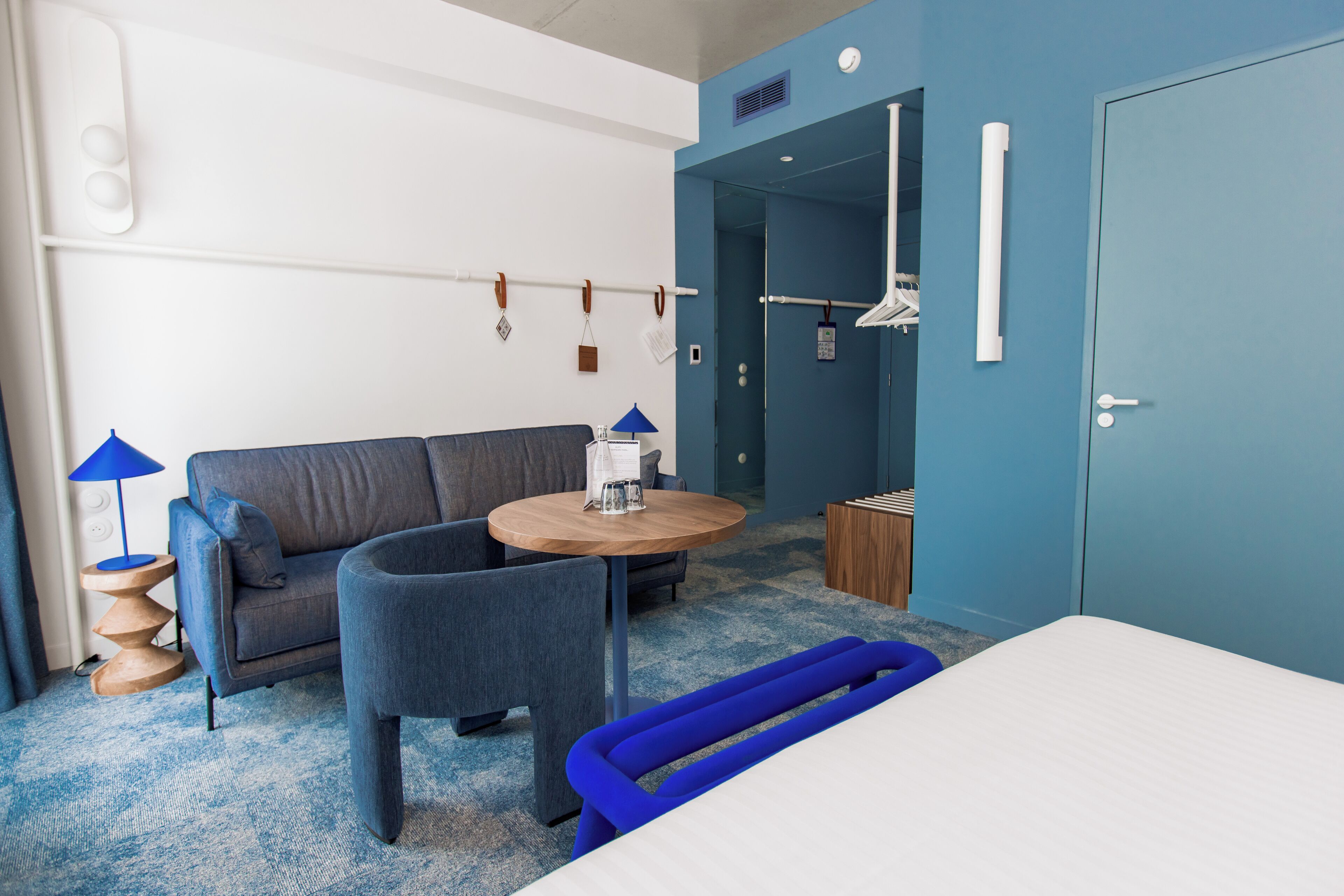 A modern hotel room with a grey sofa, a round wooden table, a blue bench, and a white bed.