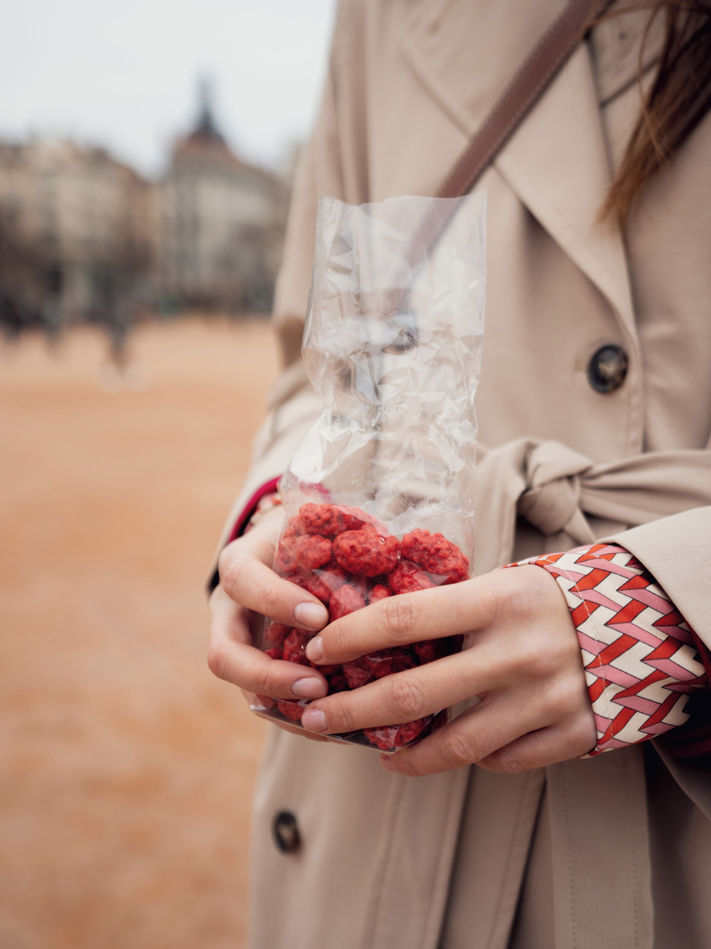 Una persona sosteniendo una bolsa de plástico transparente llena de objetos rojos, vistiendo un abrigo color beige y una camisa estampada debajo.