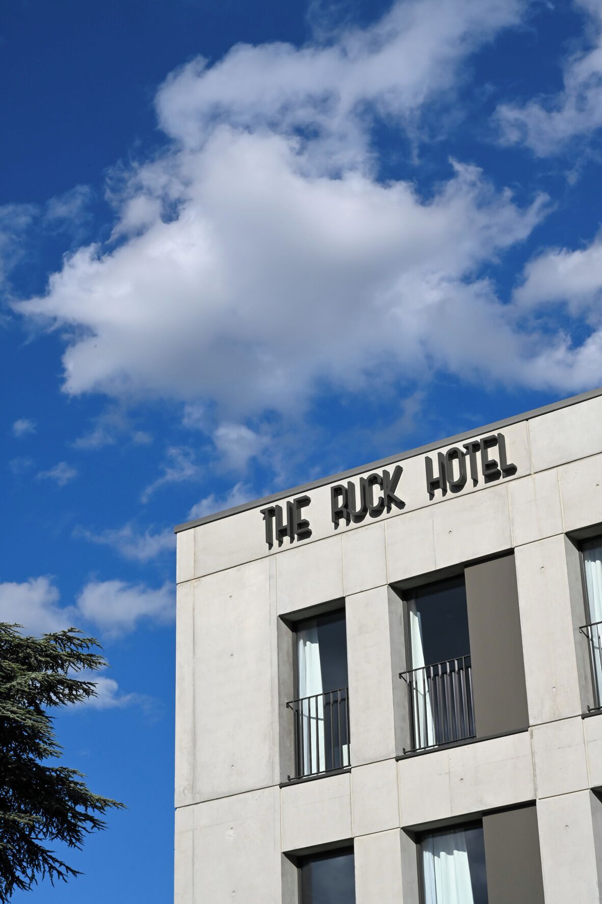 Un bâtiment portant l’enseigne « THE RUCK HOTEL », caractérisé par un style architectural moderne, avec de grandes fenêtres et un toit plat.