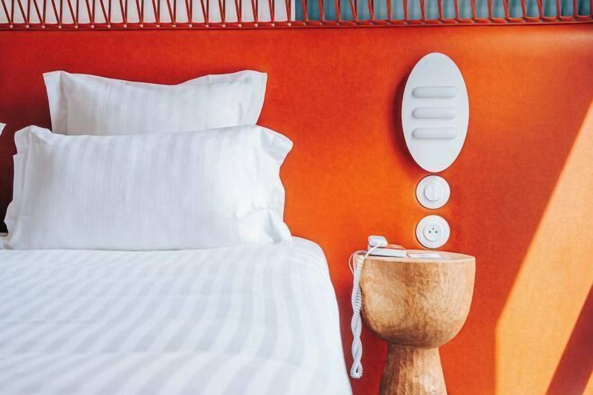Een bed met wit beddengoed, een houten nachtkastje met een telefoon en een wandlamp.