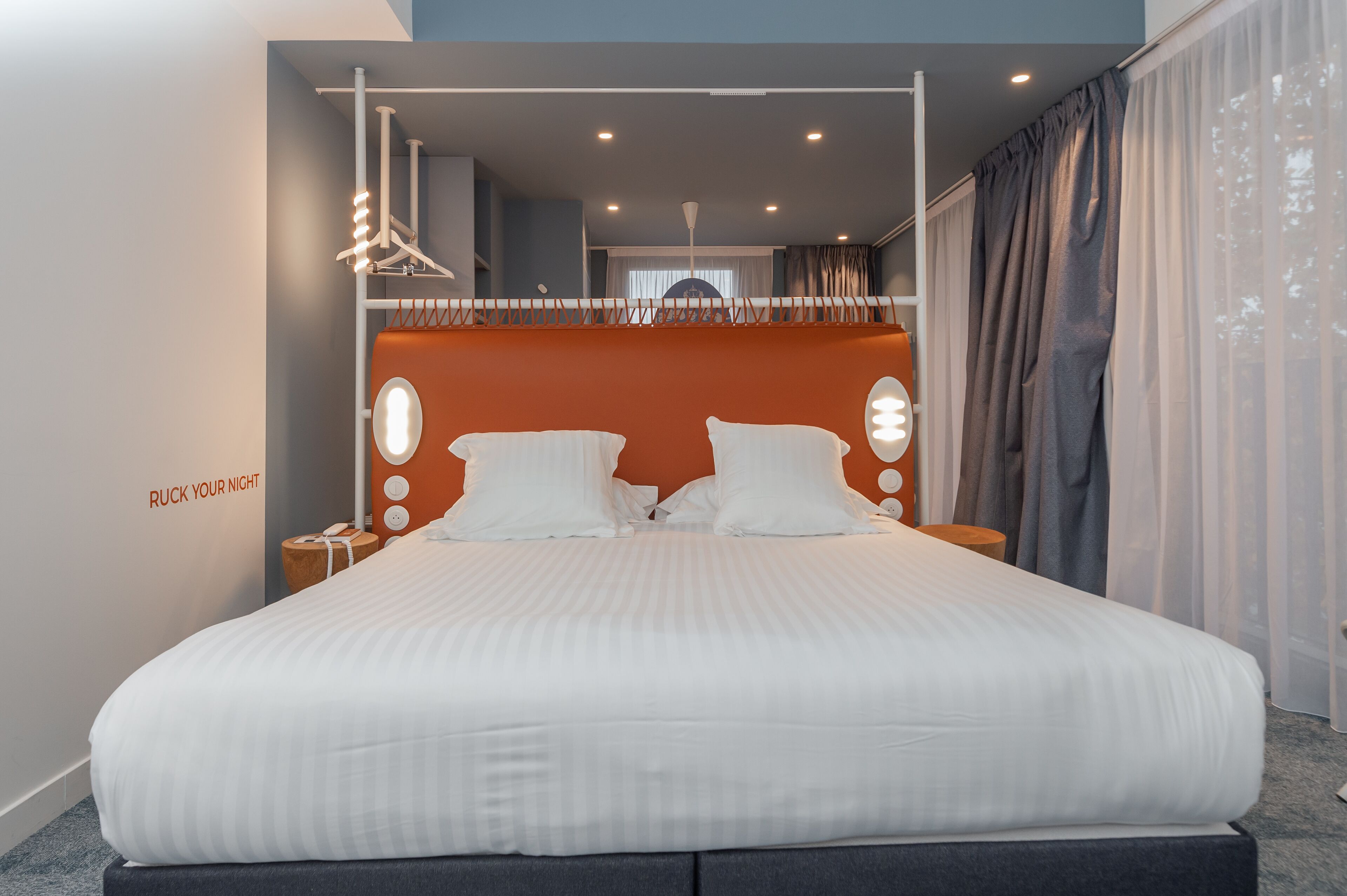 A modern hotel room with a large bed, a headboard with integrated lighting, a nightstand, and a view of the city through large windows.