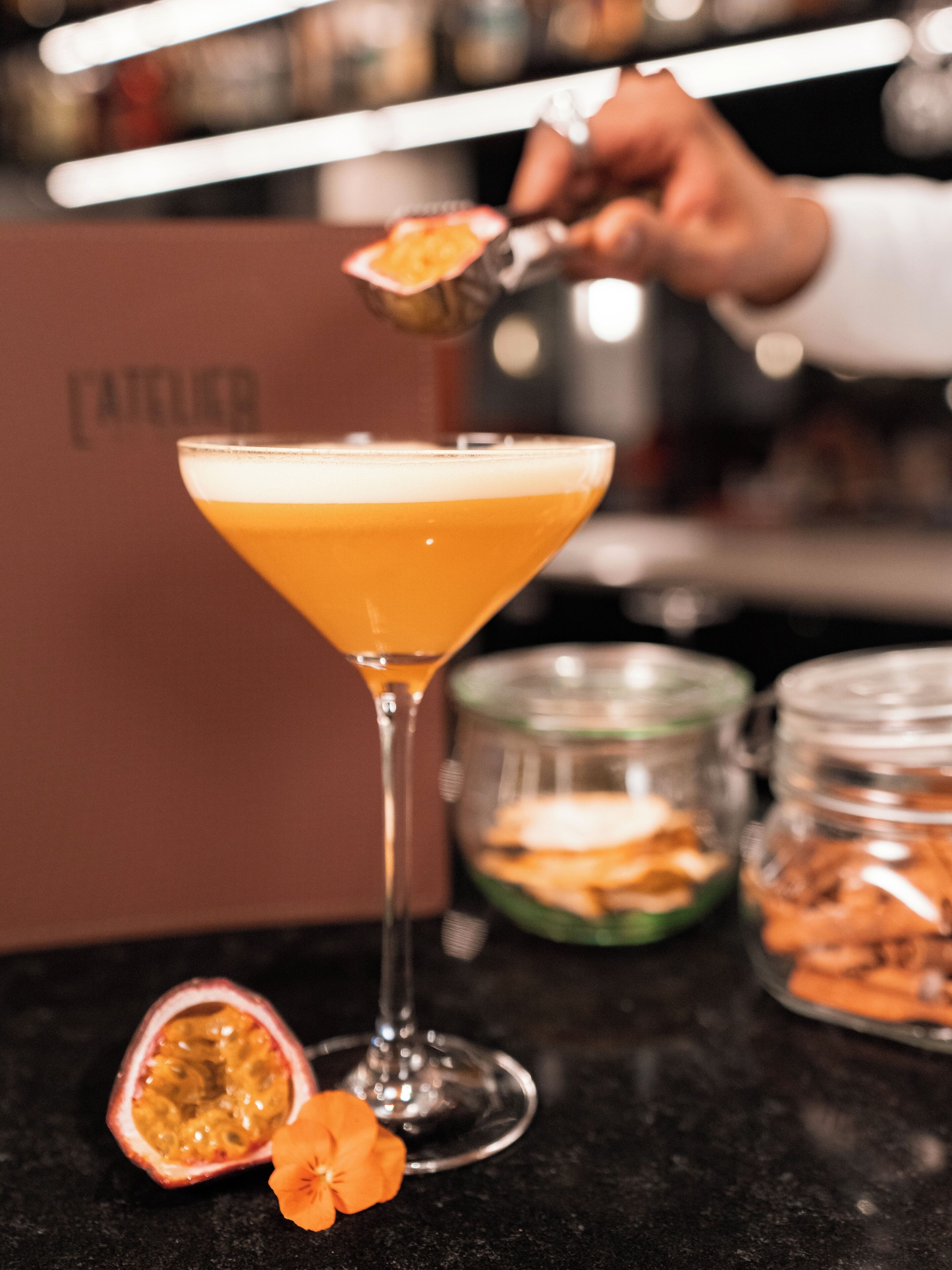 Een martini glas gevuld met een geelachtig drankje, een doormidden gesneden passievrucht en een bloem op een bar.