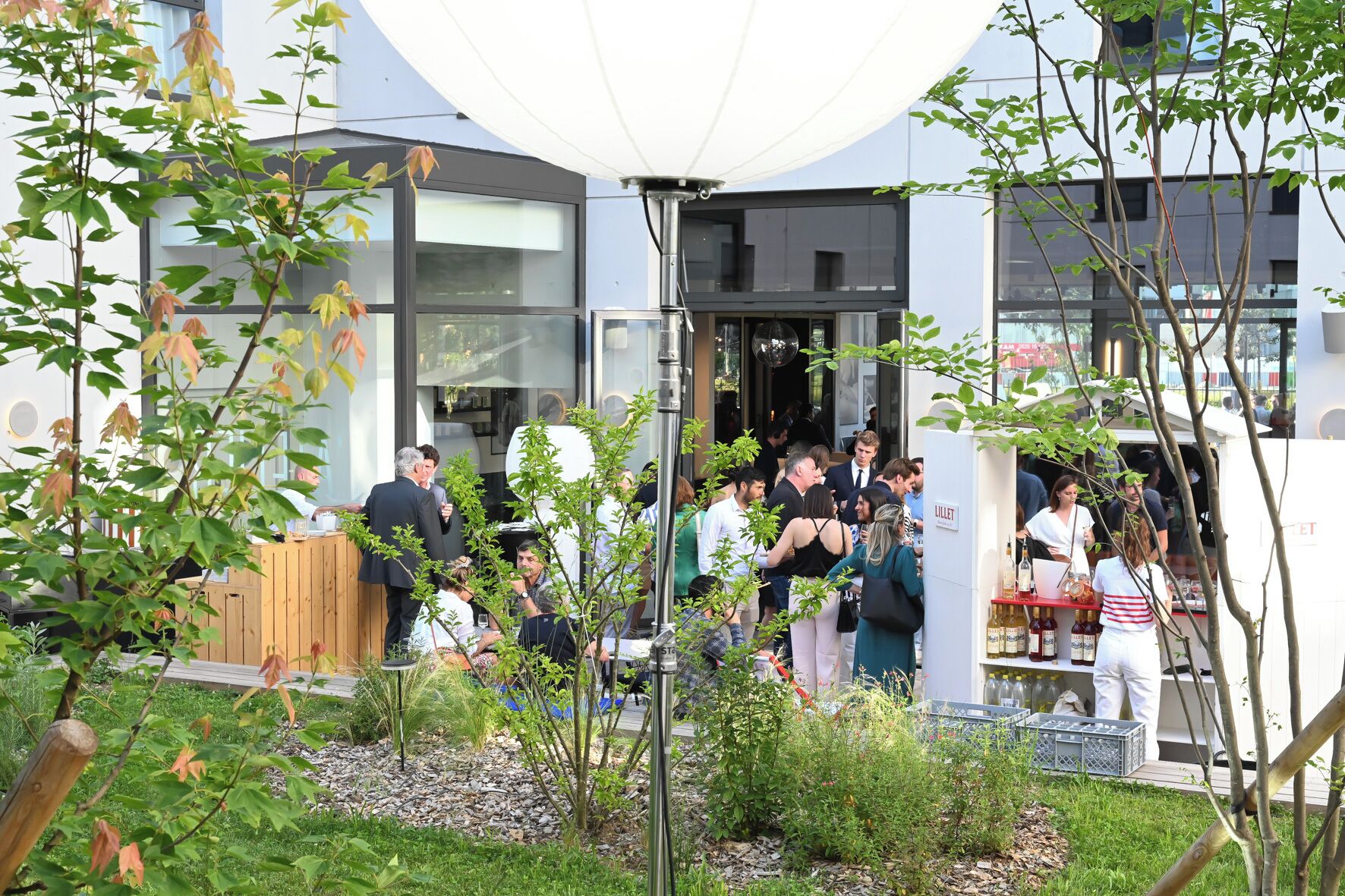 An outdoor gathering area with a modern building in the background, a wooden fence, and various plants.