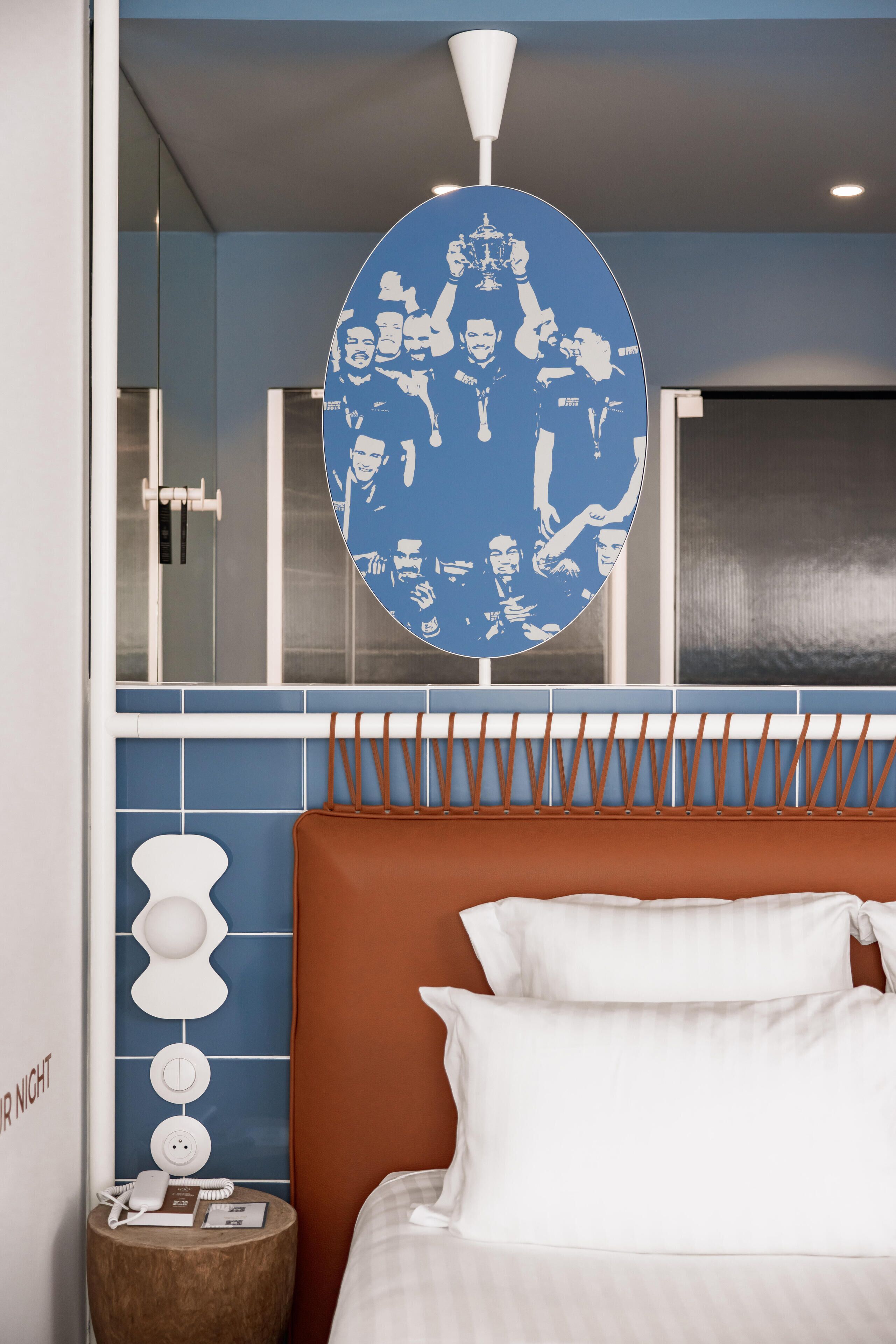 A hotel room with a large circular artwork on the wall, a bed with white linens, a brown headboard, and a bedside table with a lamp and a phone.