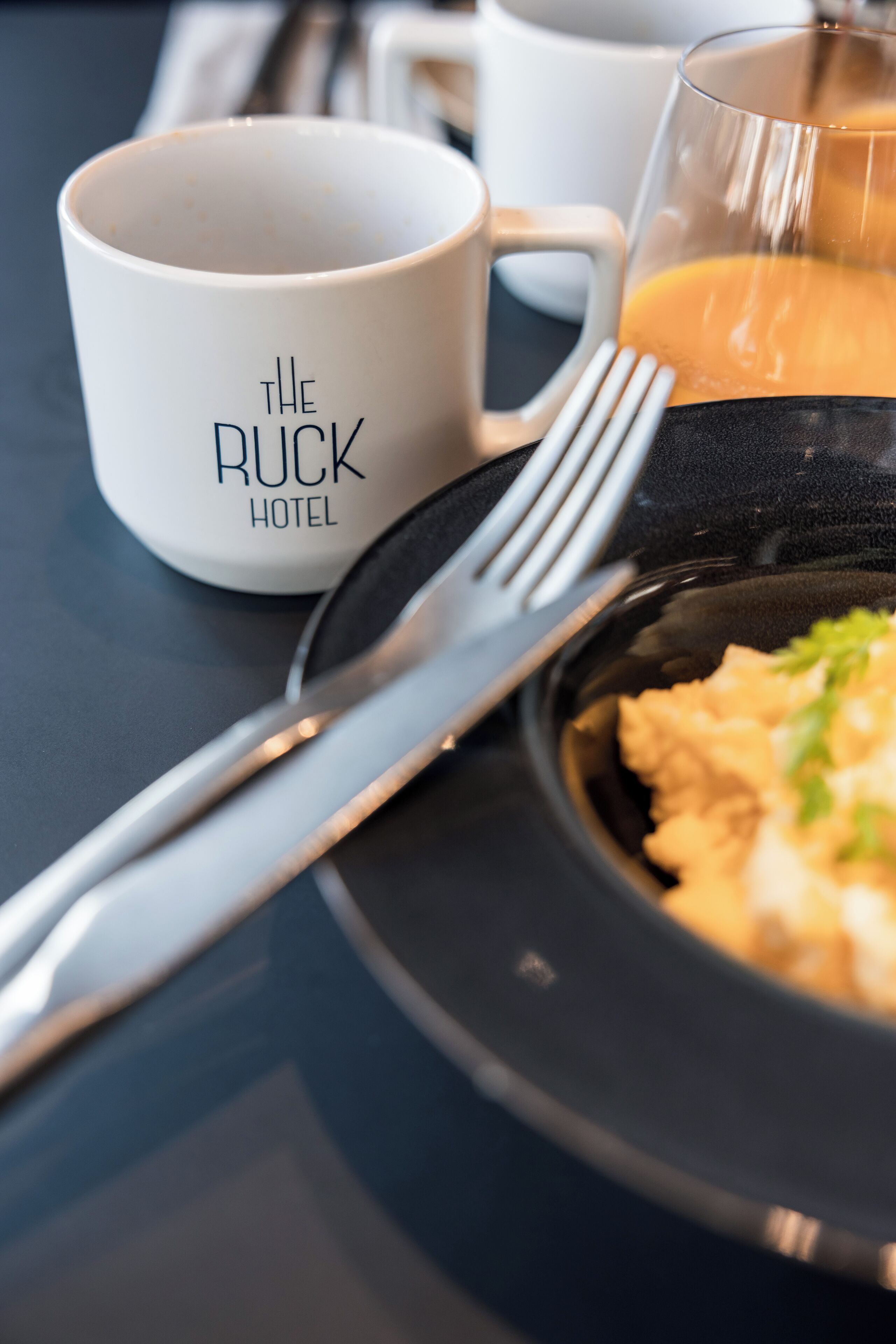 A breakfast setting with a white mug labeled "The Ruck Hotel," a fork, a glass of orange juice, and a plate with scrambled eggs.