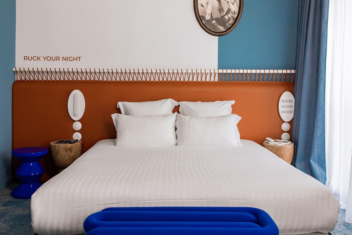 Een keurig opgemaakt bed met een blauwe bank ervoor, een wandklok en een ingelijste foto erboven.