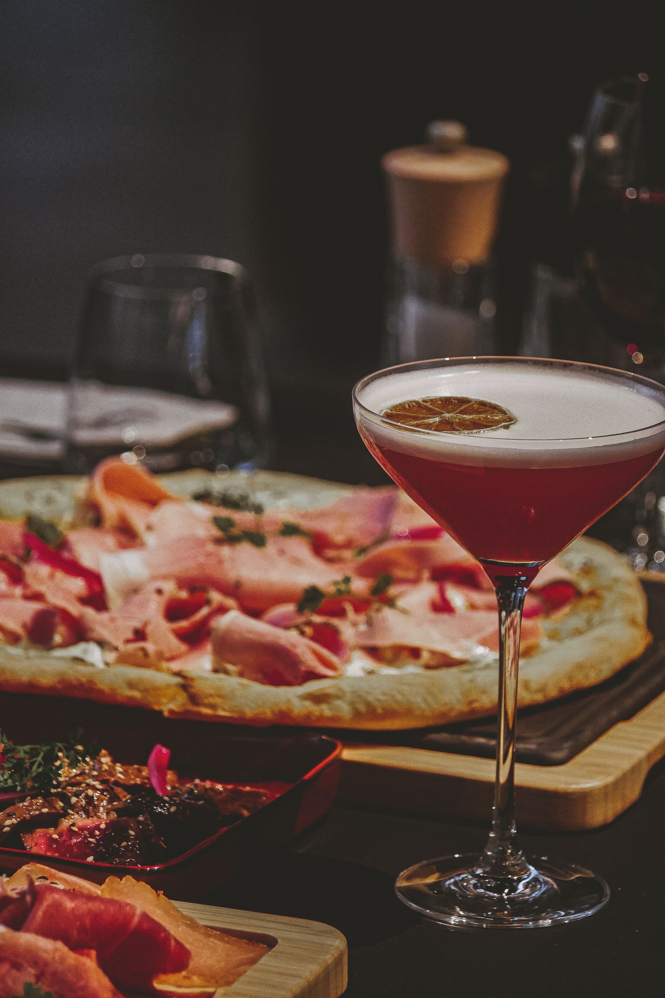 Un got de martini amb guarnició, una pizza amb diferents ingredients i un plat petit de carn.