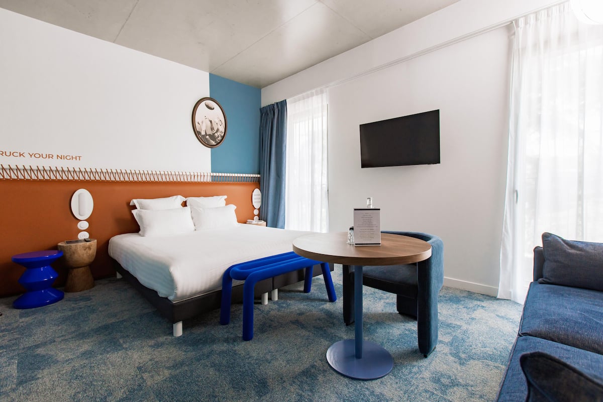 Een hotelkamer met een bed, een ronde tafel, een blauwe bank, een televisie en een muur met een klok en een bord.