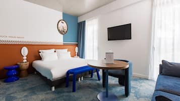 A hotel room with a bed, a round table, a blue bench, a TV, and a wall with a clock and a sign.