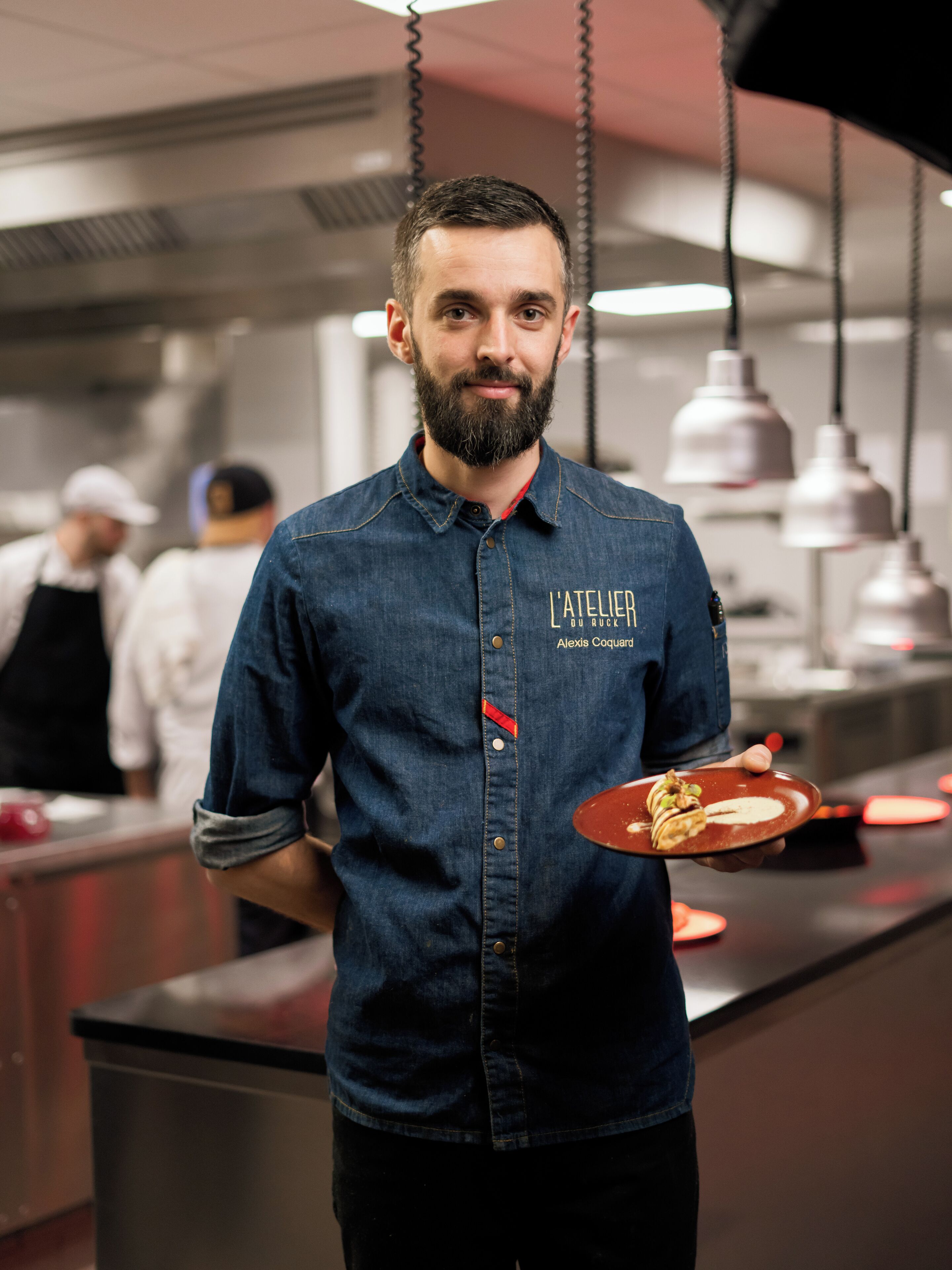 A chef in a denim shirt holding a plate with food, standing in a kitchen with other chefs in the background.
