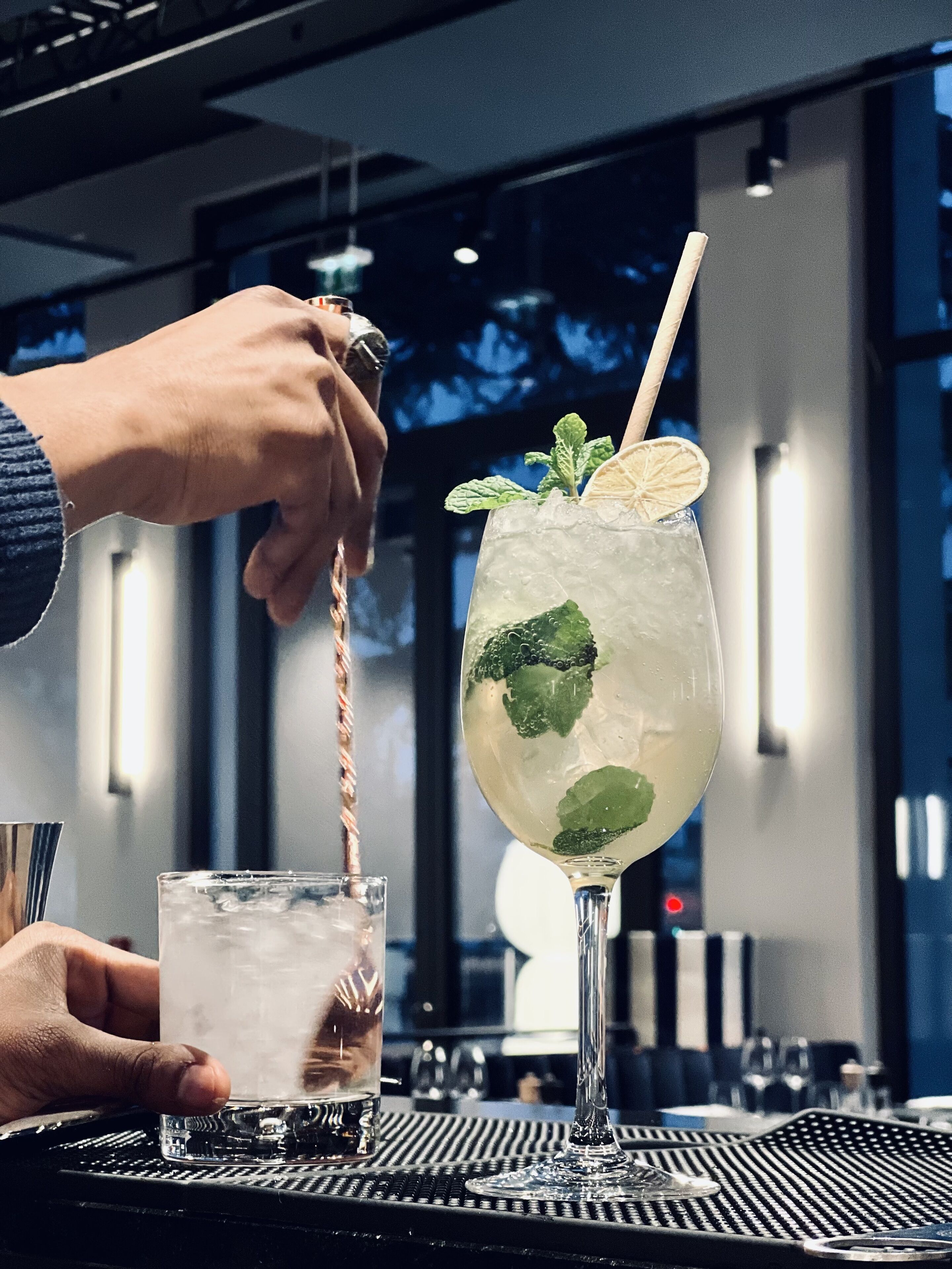 Un bar con un cantinero preparando una bebida en un vaso alto, hielo, menta y una rodaja de limón.