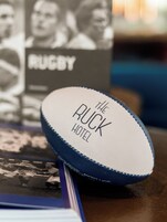 Un oggetto bianco e rotondo con la scritta "The Ruck Hotel", appoggiato su un tavolo con uno sfondo sfocato che include un elemento a tema rugby.