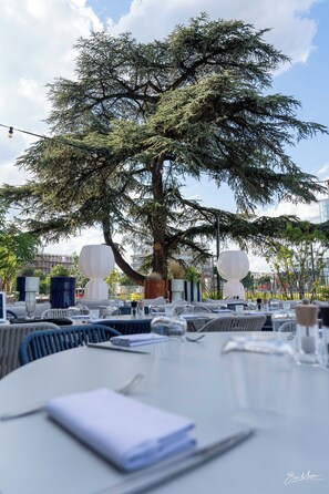 Un área de comedor al aire libre con mesas y sillas, un árbol grande y vistas de edificios al fondo.