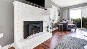 A living room with a white brick fireplace, a flat-screen TV mounted above it, a grey sofa, and a large window offering a view of the outdoors.