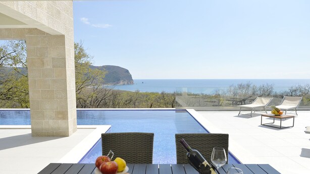 Ein Esstisch mit Obst und Weingläsern, mit Blick auf einen Pool und eine malerische Meerlandschaft.