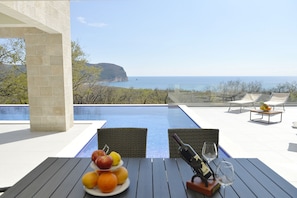 A dining table with fruit and wine glasses, overlooking a pool and a scenic ocean view.