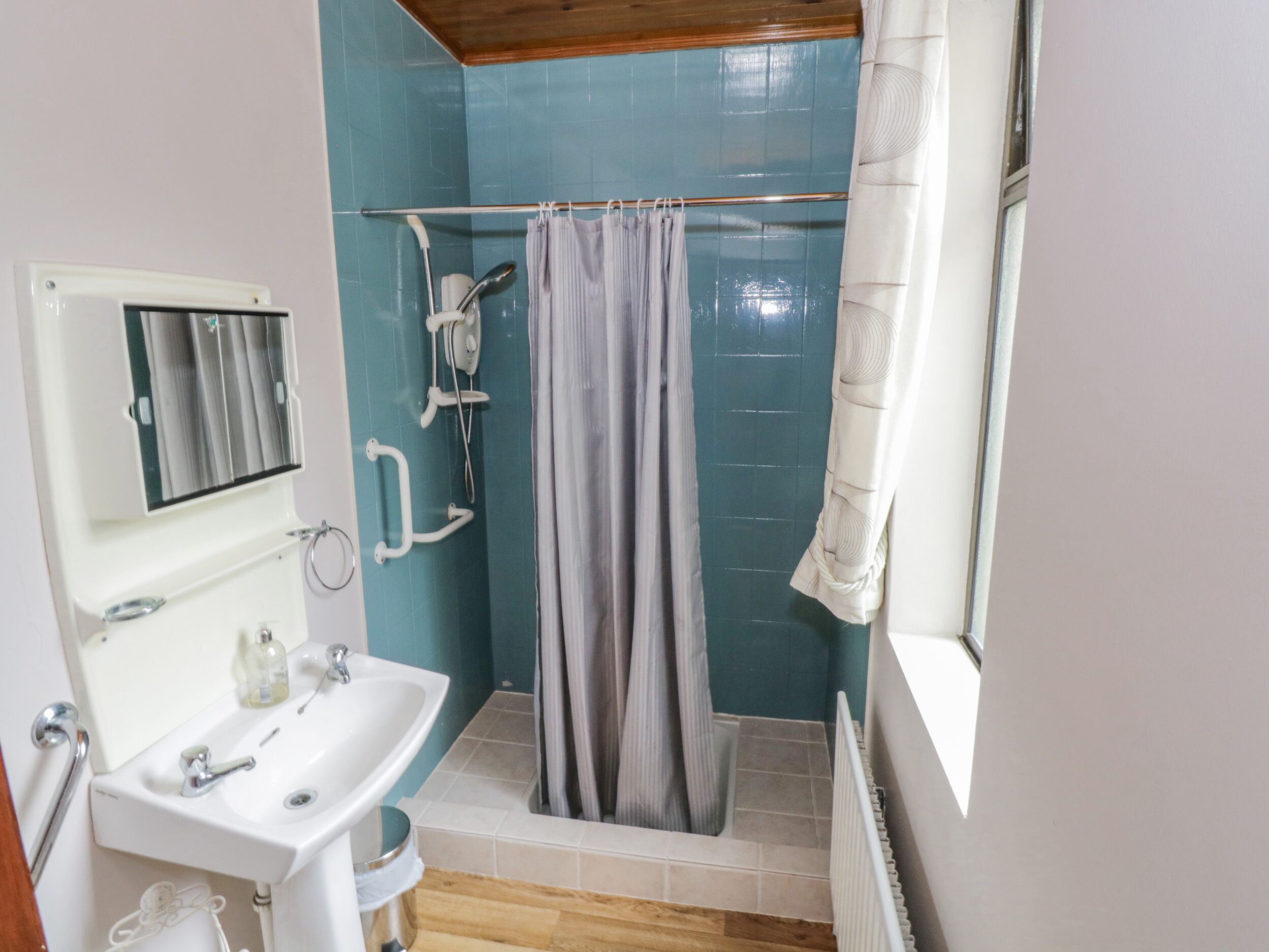 A bathroom with a sink, mirror, shower, and a window with a curtain.