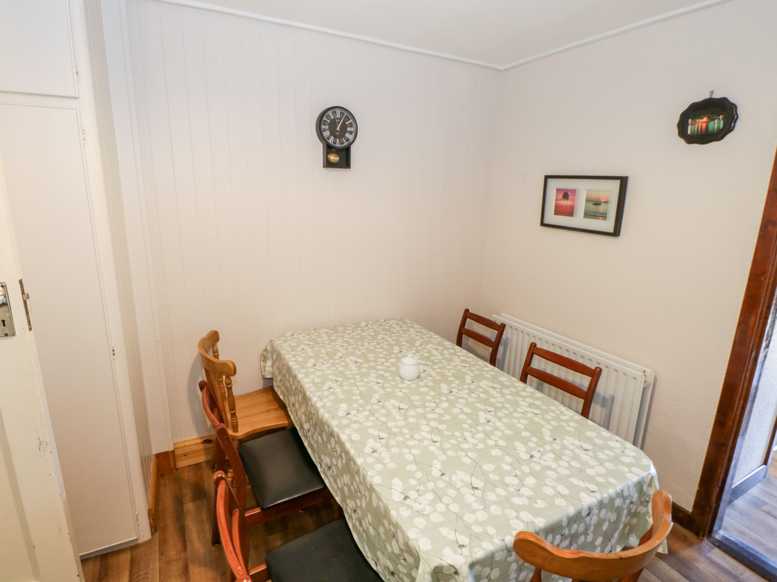 Uma pequena sala de jantar com uma mesa coberta por toalha florida, duas cadeiras de madeira, um relógio de parede, um quadro emoldurado e um radiador.