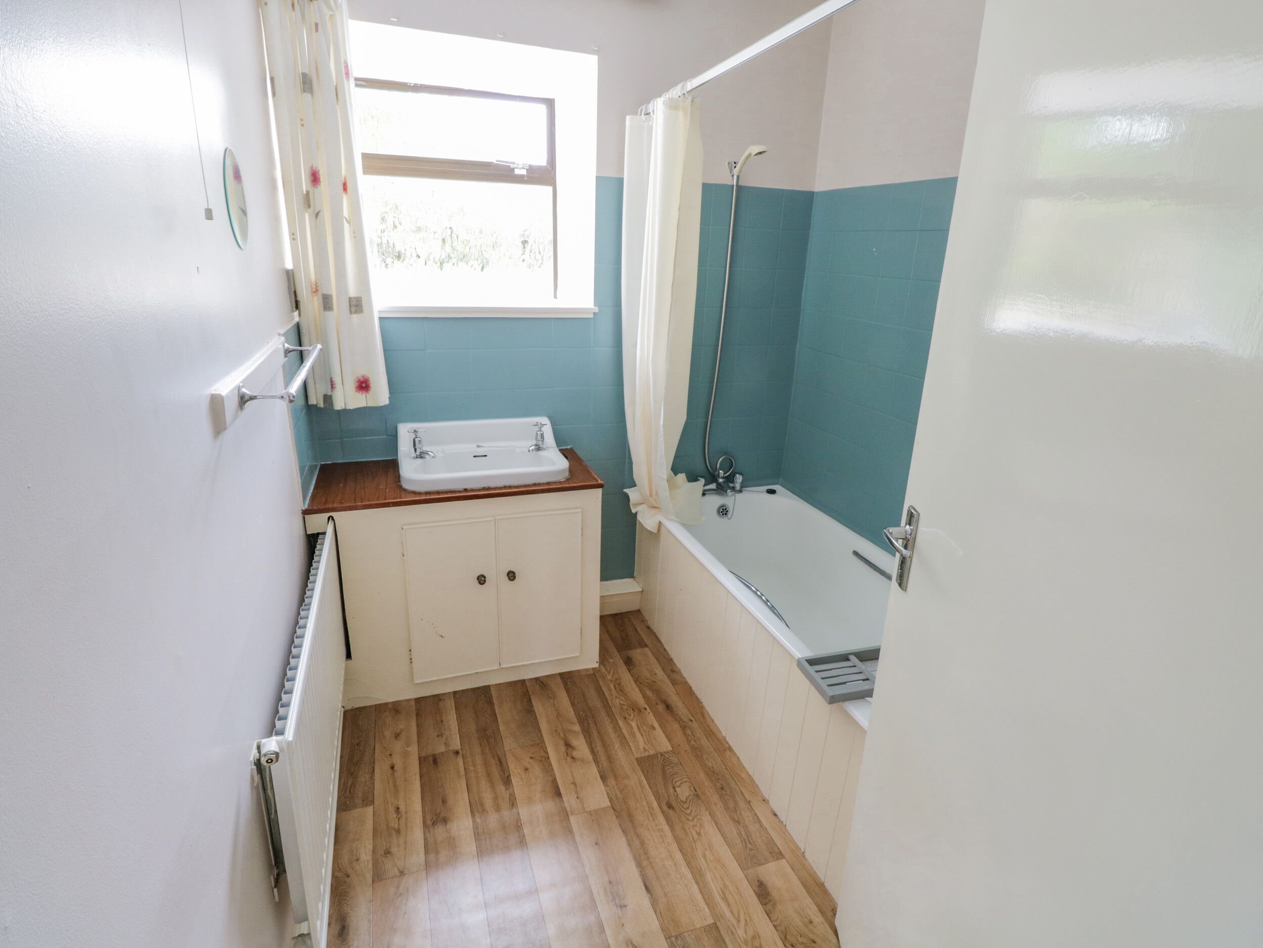 Un baño pequeño con piso de madera, un lavamanos blanco, una bañera y una ventana con cortinas.