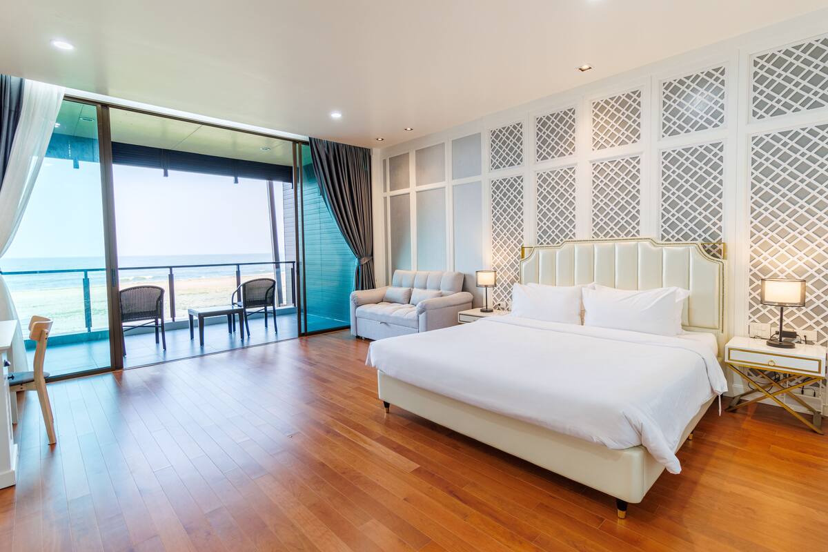 A spacious bedroom with a large bed, a seating area with chairs, a view of the sea through a glass door, and a patterned wall.