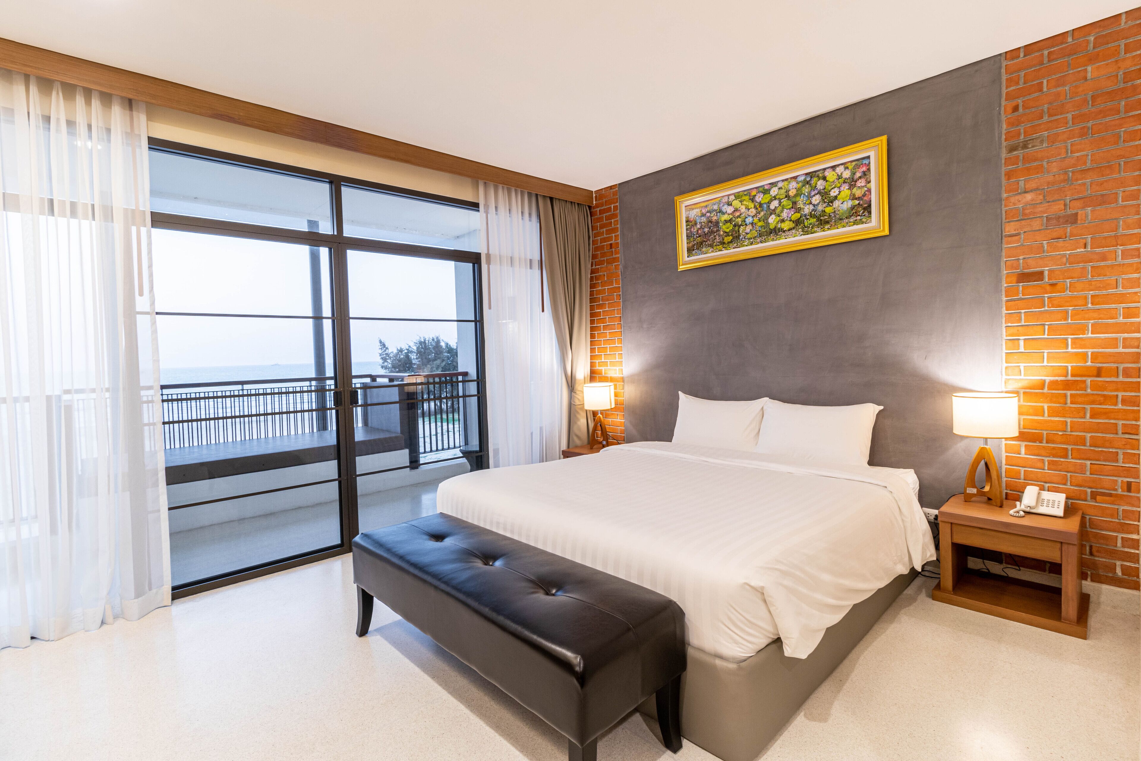 A hotel room with a large bed, a bench, a nightstand with a phone, a window with curtains, and a view of the outdoors.
