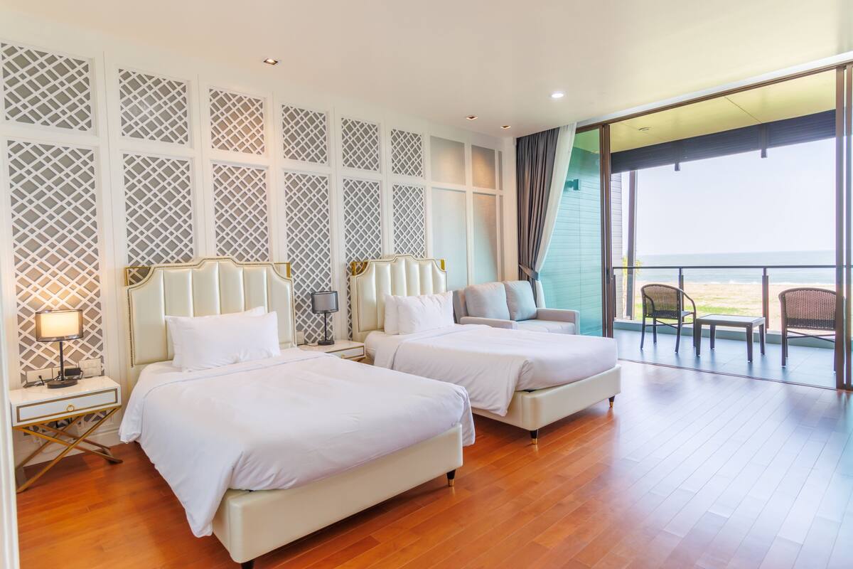 A hotel room with two beds, a balcony with a view of the beach, and a large window.