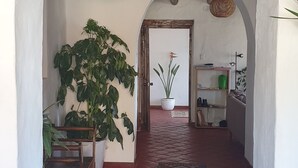 A hallway with a red-tiled floor, white walls, and wooden beams on the ceiling. There are two hanging wicker lamps, a potted plant, and a chair. - Ecolodge and Retreat "Casa de Colibri (Cajamarca)