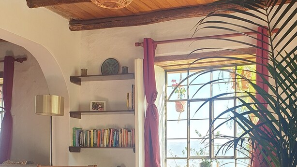 A cozy living room with a wooden ceiling, a bookshelf, a sofa, and a large window with curtains. - Ecolodge and Retreat "Casa de Colibri (Cajamarca)