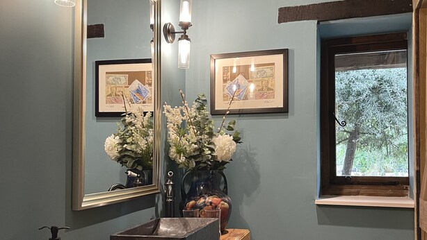 A bathroom with a wooden vanity, a large mirror, wall-mounted lights, framed artwork, and a window with a view of trees.