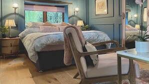 A bedroom with a large bed, a chair, a desk, a mirror, and a window with a view of greenery.