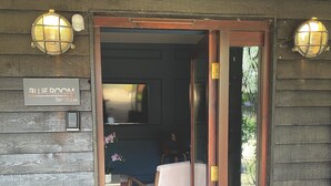 A wooden door leading to a room with a bed, a chair, a table, and a TV. There are two wall-mounted lights and a sign above the door reading "Blue Room."