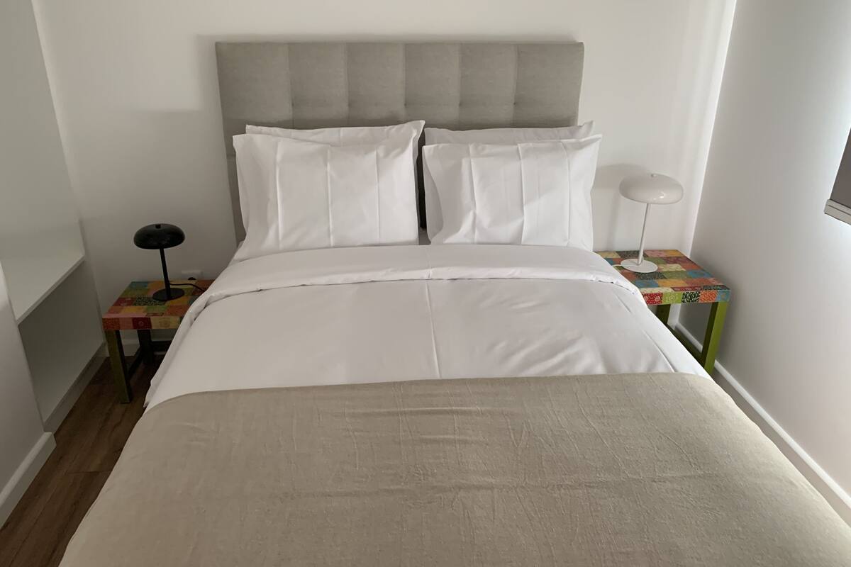 Un lit bien fait, avec du linge de lit blanc, une tête de lit rembourrée de couleur grise et une petite table de chevet sur laquelle se trouvent une lampe et une nappe colorée à motifs matelassés.