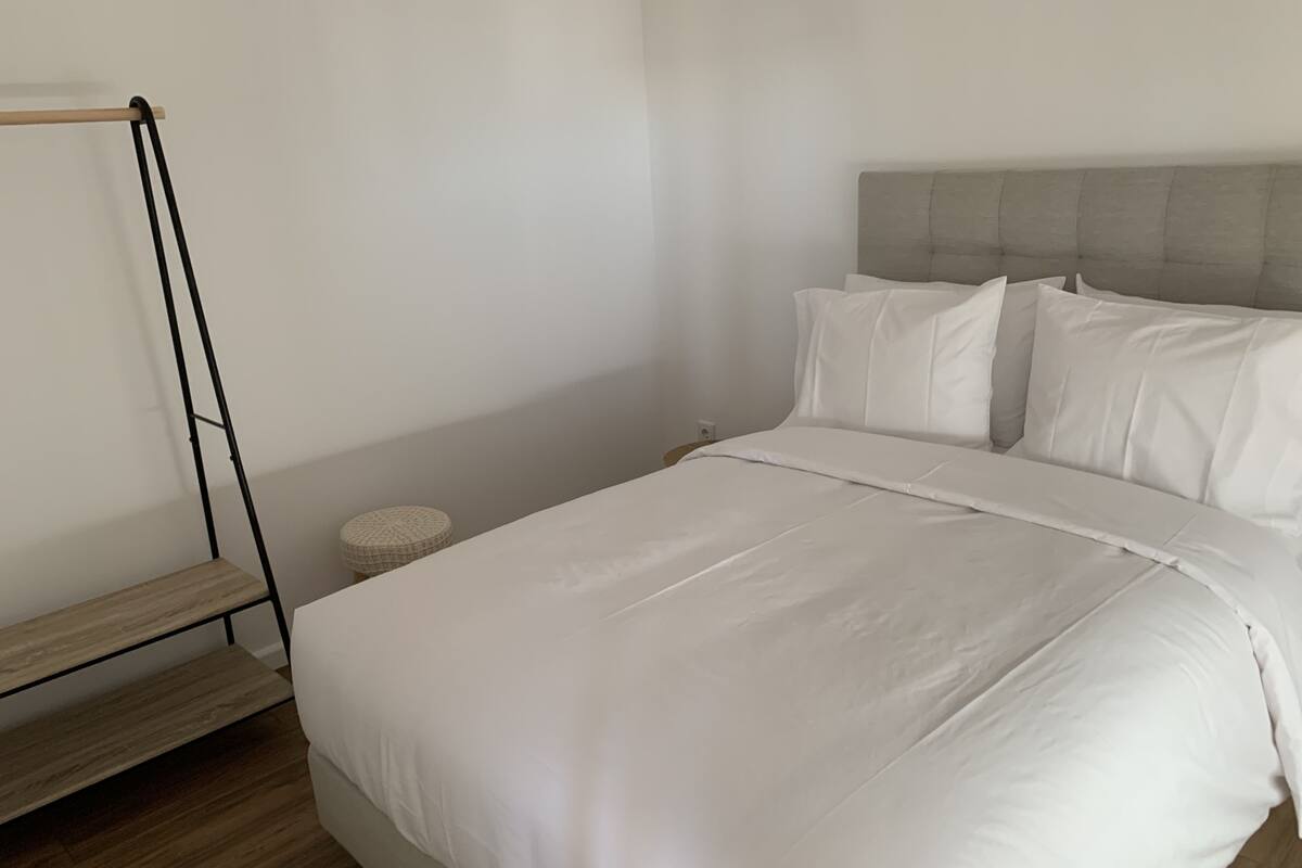 Un lit bien fait, avec du linge de lit blanc, une tête de lit rembourrée grise et un petit pouf rond placé à côté.