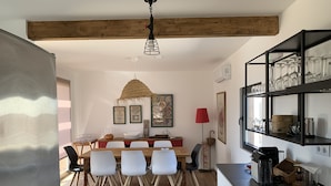A dining area with a wooden table and chairs, a hanging light fixture, and a shelf with glassware.