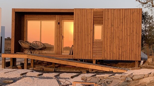 A modern wooden house with a deck, outdoor seating, and a large glass door.