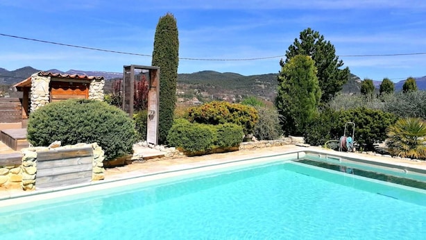 Una piscina amb terrassa de pedra, envoltada d'arbustos i arbres diversos, i una petita construcció de fusta amb un mur de pedra.