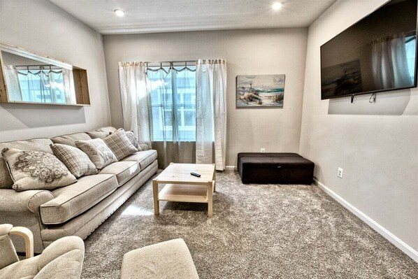 A living room with a sofa, a coffee table, a black ottoman, a TV mounted on the wall, and a window with curtains.
