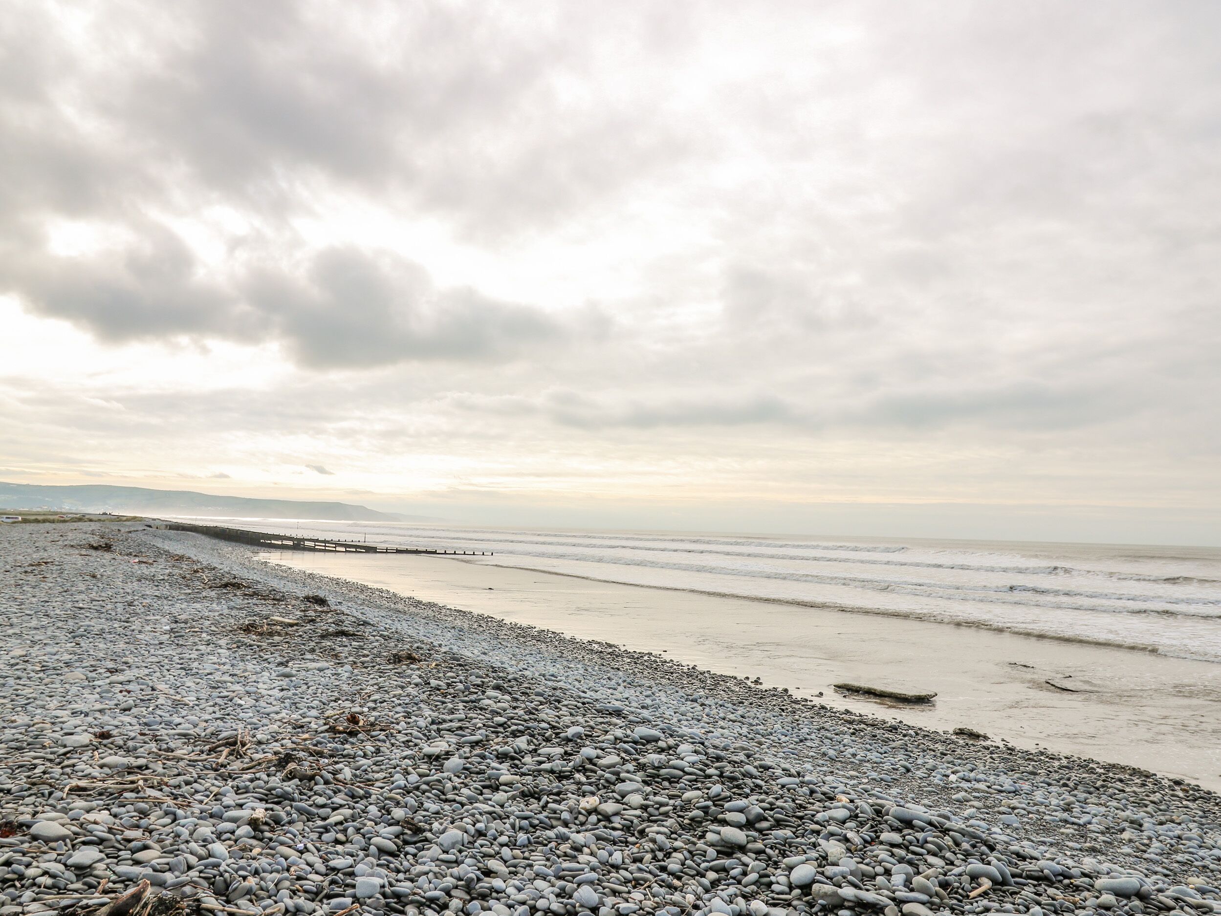 A pebbled beach with a wooden pier extending into calm waters under a cloudy sky.