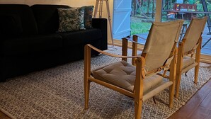 Une pièce chaleureuse avec un canapé aux couleurs sombres, un plancher en bois, un tapis tissé et deux chaises en bois dotées d’assises en toile.