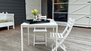 Une table et des chaises blanches sont disposées sur une terrasse en bois, avec un banc blanc et une porte blanche en arrière-plan.