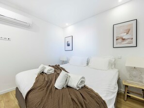 A hotel room with a bed, a brown blanket, white pillows, a framed picture on the wall, and an air conditioning unit.
