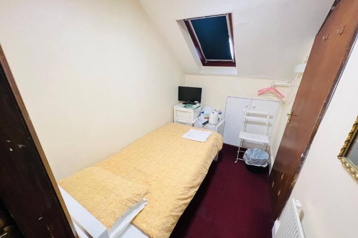 A small, narrow room with a bed, a desk, a chair, a TV, a radiator, and a window.