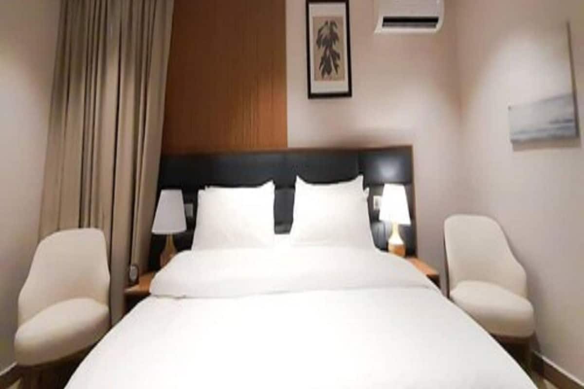 A hotel room with a large bed, two bedside tables with lamps, a headboard, an air conditioning unit, a window with curtains, and a framed picture on the wall.