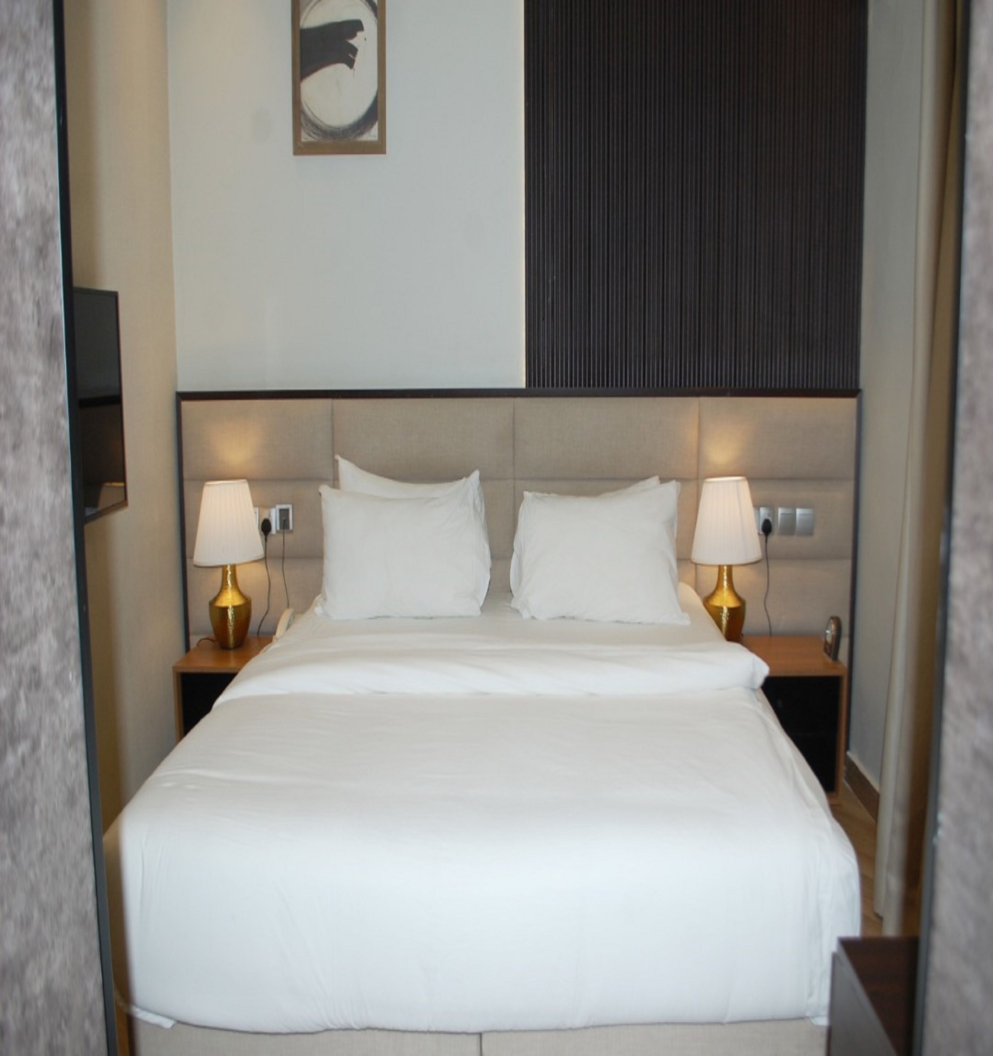 A hotel room with a large bed, two bedside tables with lamps, a wall-mounted TV, and a large abstract artwork above the bed.