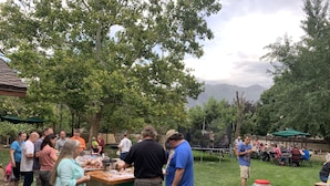An outdoor gathering with people around food and drink stations, tables, and a grassy area with trees.