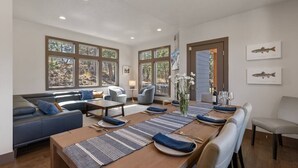 Un espace de salle à manger moderne, avec une longue table en bois dressée pour six personnes, flanquée de canapés bleus et d’un coin salon comprenant un canapé et des fauteuils.