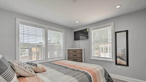 Quarto com uma cama grande, duas janelas com persianas, uma TV fixada na parede e uma cômoda de madeira.