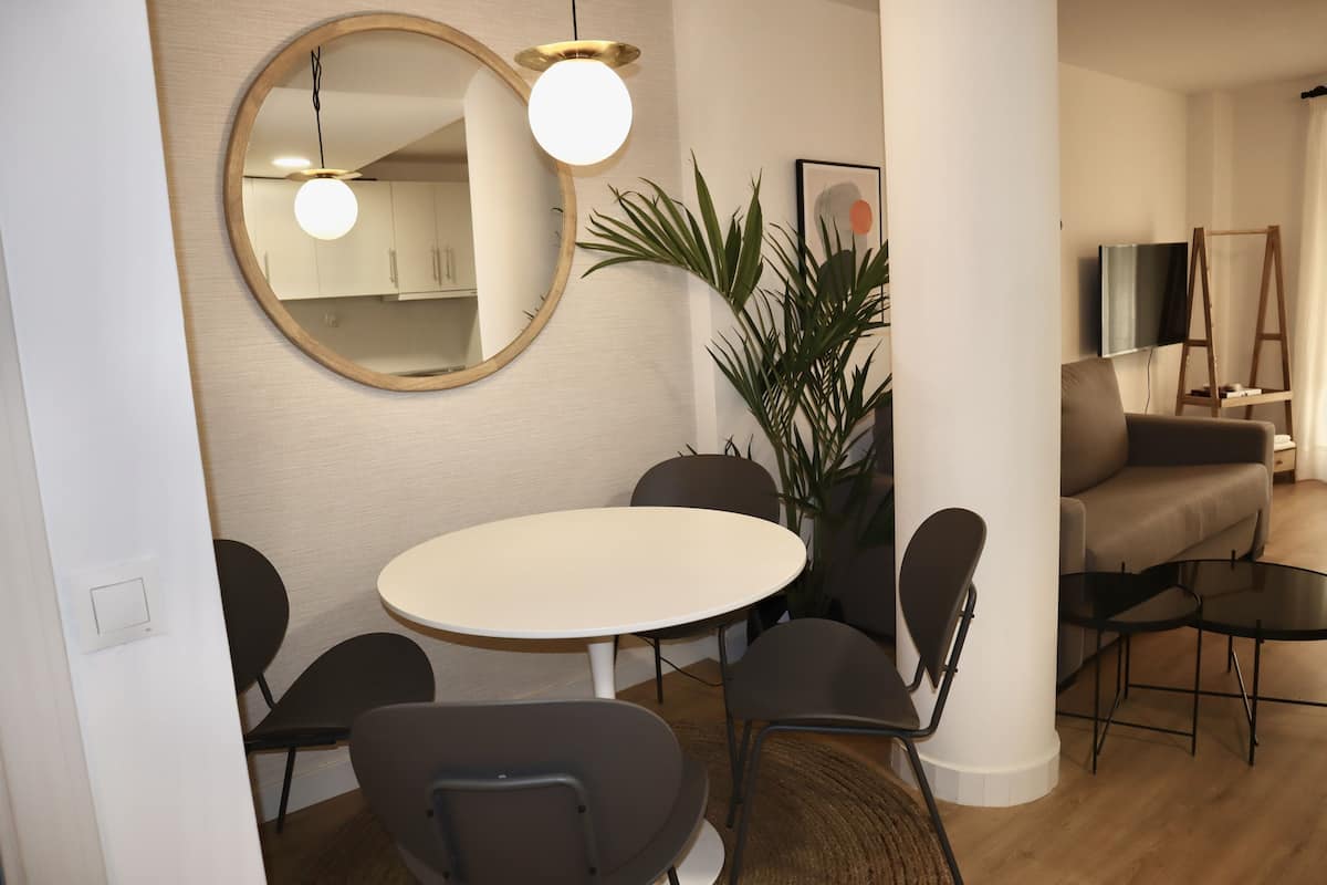 Un salon moderne avec une table à manger ronde, six chaises, un grand miroir, un téléviseur, un canapé et une plante.