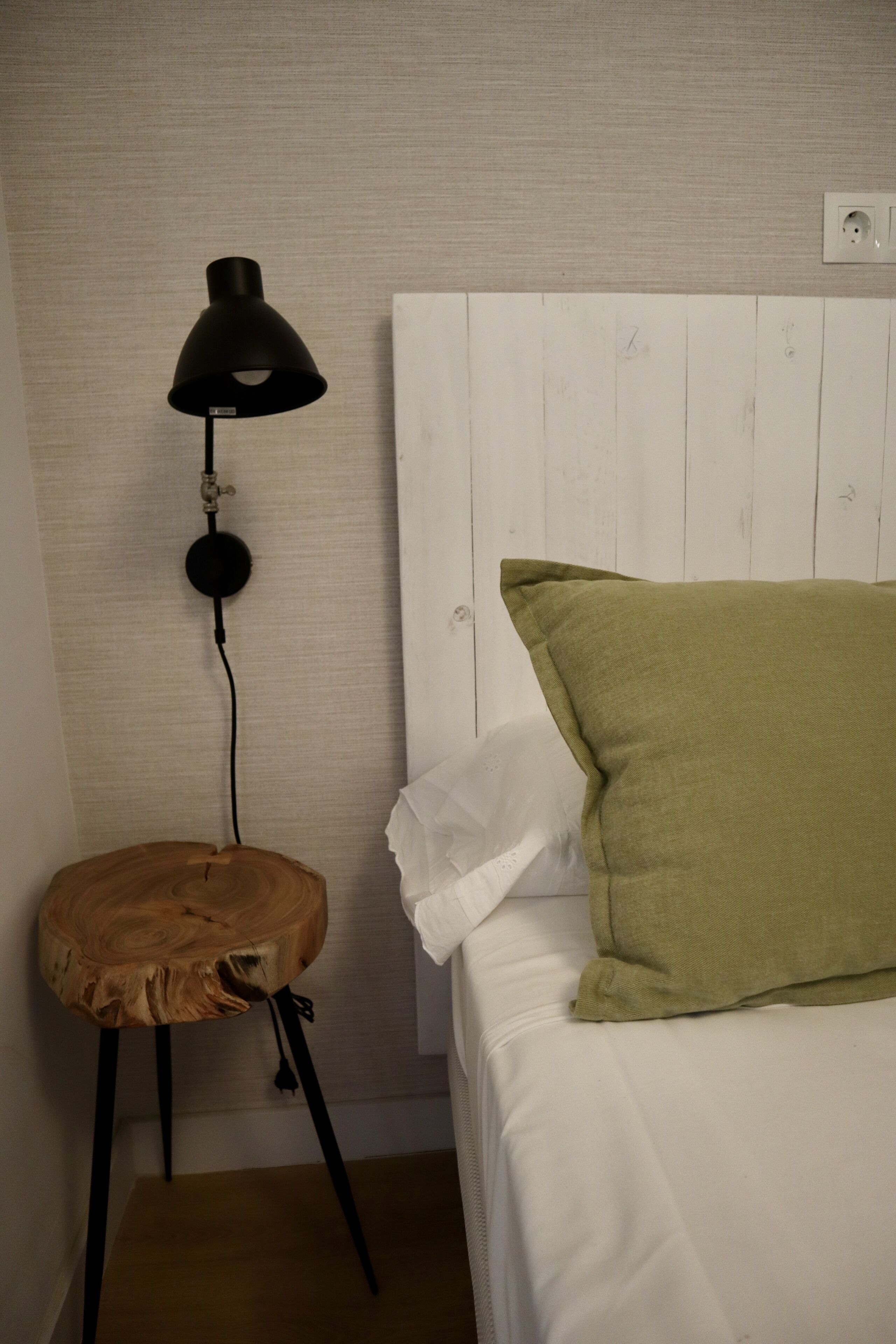 Une pièce avec un lit blanc, un oreiller vert, un oreiller blanc, une lampe noire et un tabouret en bois.