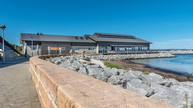 A modern building with a flat roof, large windows, and a stone retaining wall by the water.